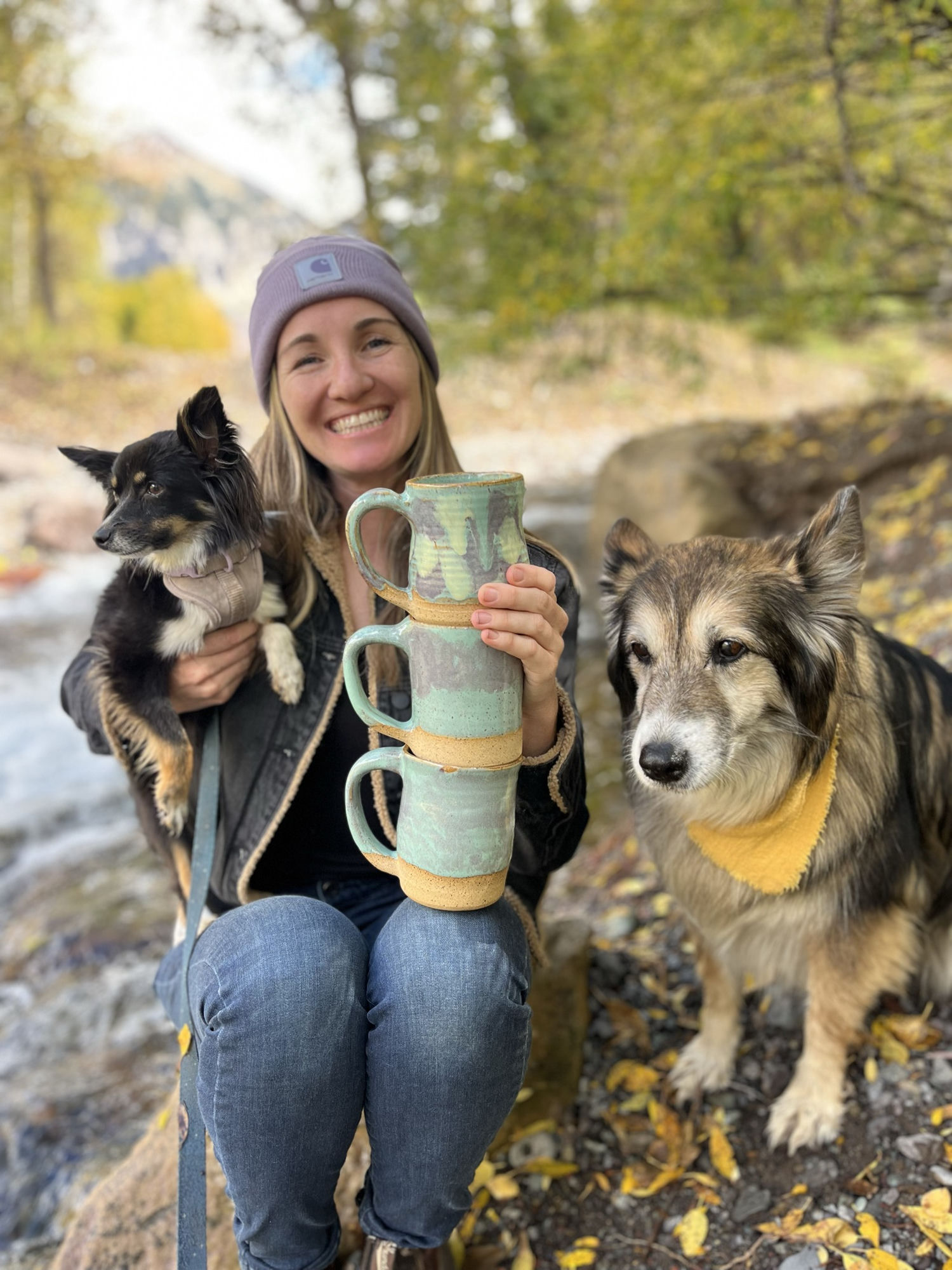 Nancy with her two dogs, holding a stack of her mugs beside a mountain river
